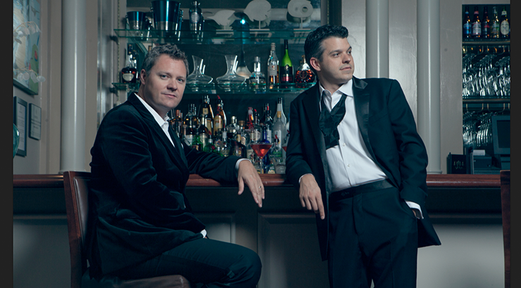Two men in suits, leaning on a bar, one sitting on a barstool and the other standing beside him 