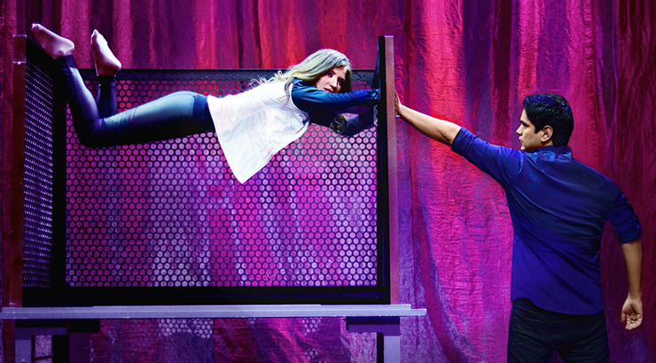 Jason Bishop on stage, placing his hand on a cage-style box where a female assistant appears to be lying in the air or levitating 