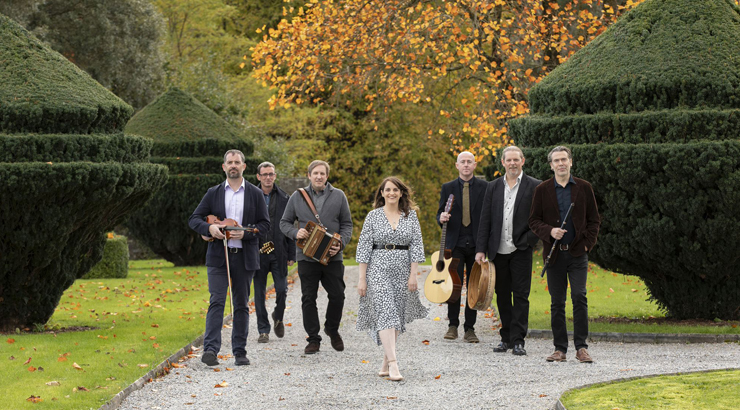 members of Danu holding various musical instruments, while walking among large green bushes and trees with fall foliage.