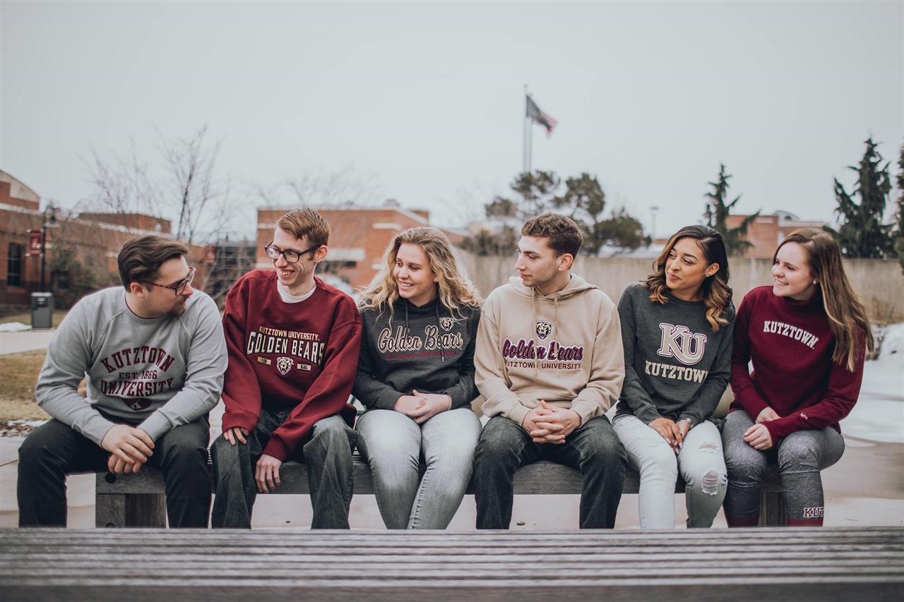 Six students sitting on a bench outside, wearing various Kutztown University apparel 