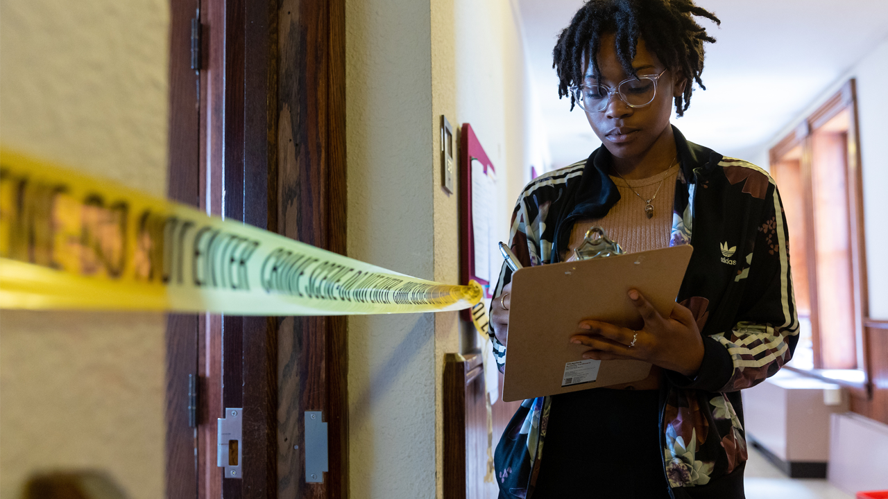 Student takes notes on a clipboard outside of a room with police tape across the entry as part of a mock crime scene learning experience. 