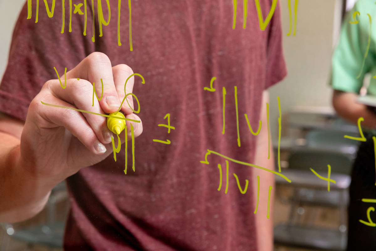 Student writing mathematic equation in yellow marker on a pane of glass