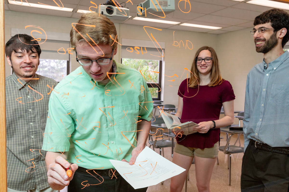 Student in the foreground copies an equation from the notes in his hand while others in the background watch him and smile