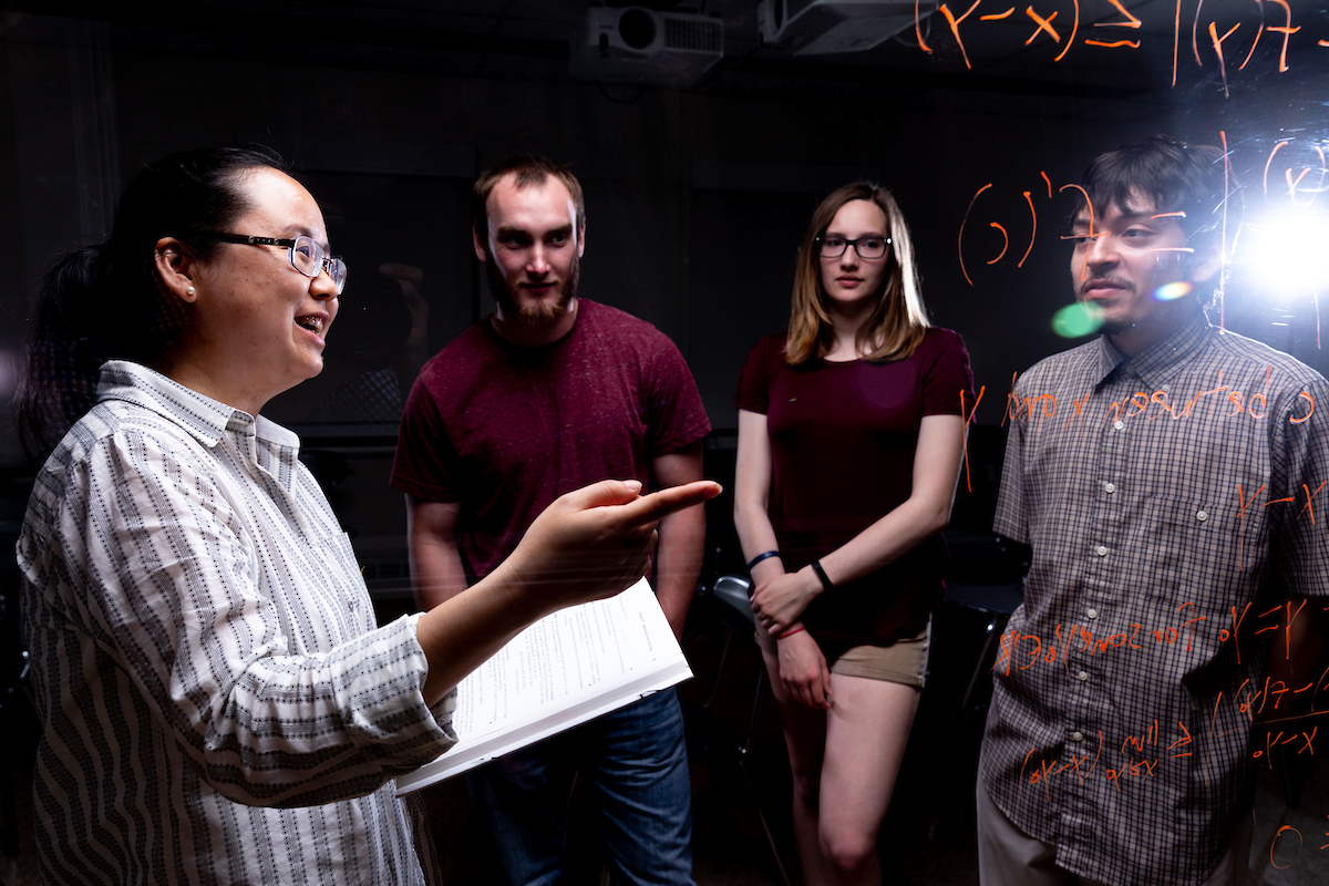 Professor explaining an equation to students in the background, pointing to the pane of glass where he wrote it in the foreground