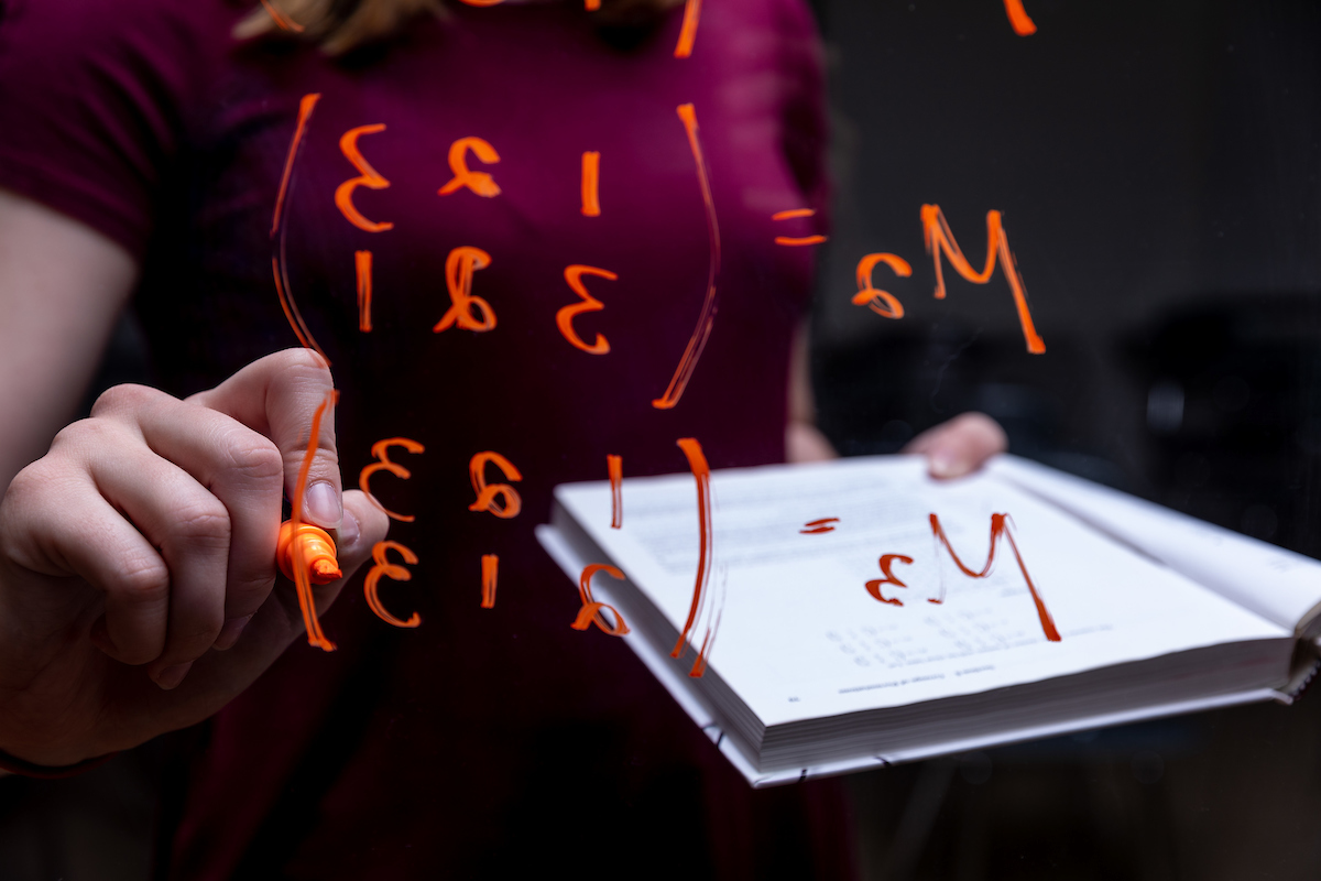 Student writing an equation an open textbook in the background on a pane of glass in the foreground