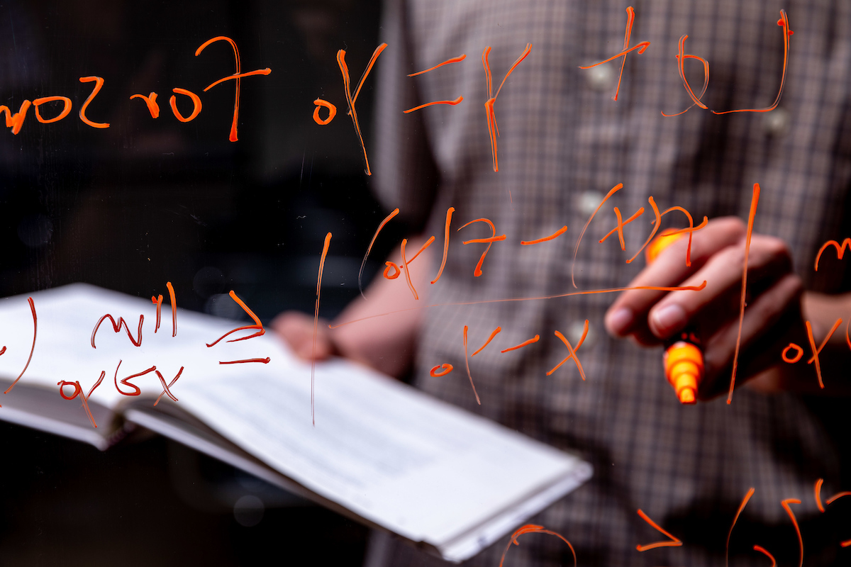 Student writing an equation from a math textbook on a pane of glass in orange marker