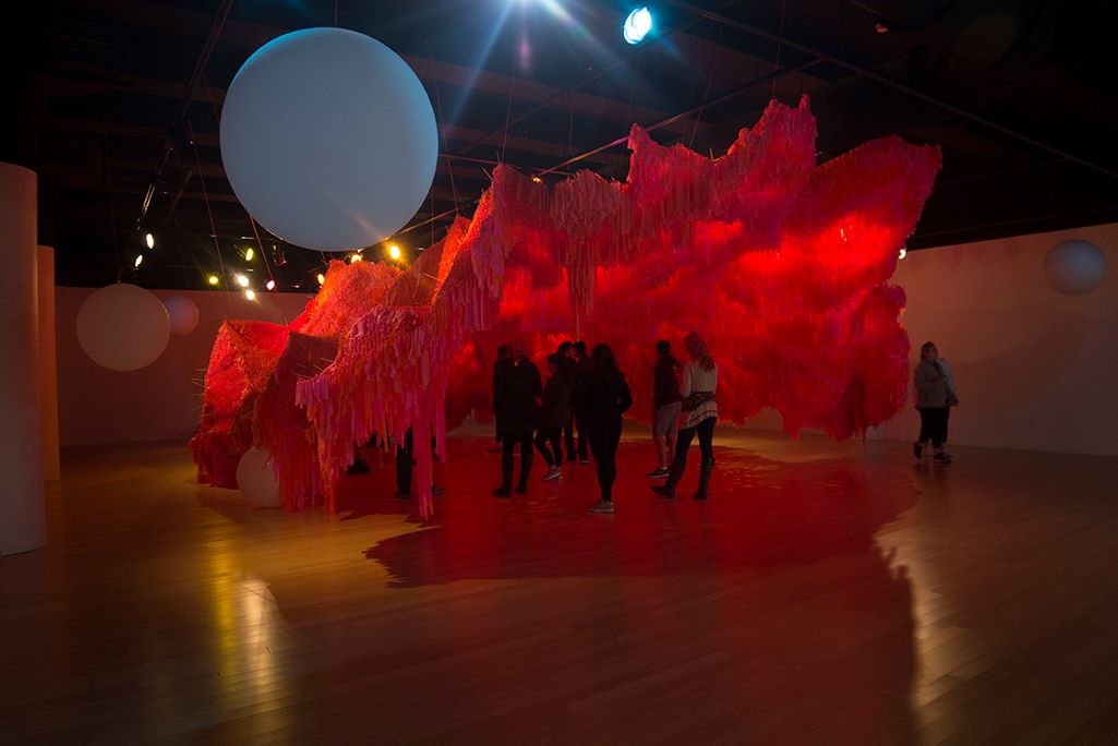 Group of observers walking under an instillation that appears to be a shapeless, glowing red cave