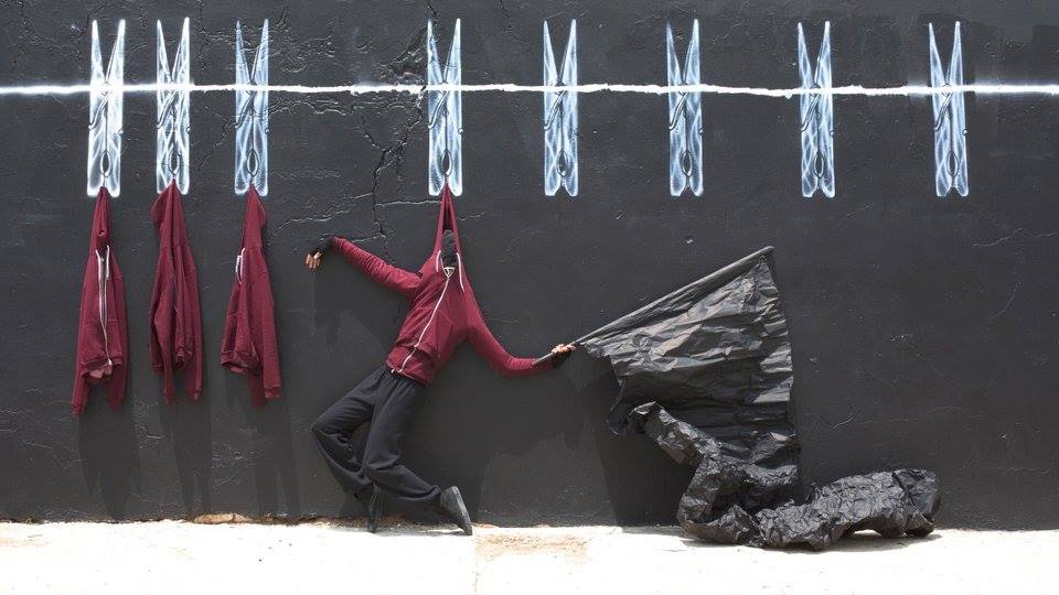 Chalk drawing of a clothesline and clothespins on a blackboard wall, with real sweatshirts and a mannequin wearing a similar sweatshirt appearing to hang from the drawn clothespins 