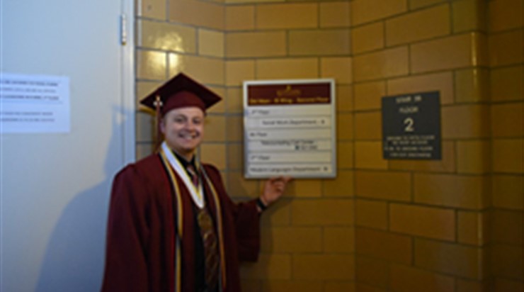 Justin Ungaro smiling in graduation robes and gesturing to a floor sign in Old Main 