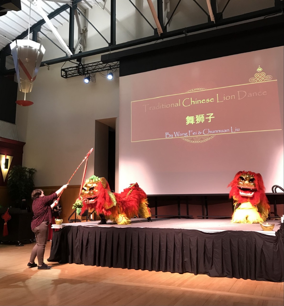Female presenter pointing with a large stick toward a projection screen with a slide that says "Traditional Chinese lion dance" in English and Chinese 