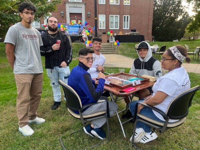 4 students sitting and 2 standing outside playing dominoes at a small table on the lawn.