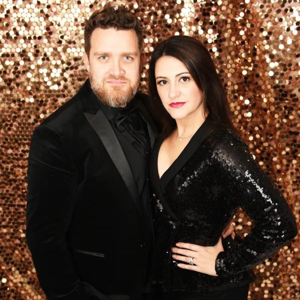 Image of man in dark suit with arm around woman on right - both dressed in shiny, black clothing with shiny gold backround