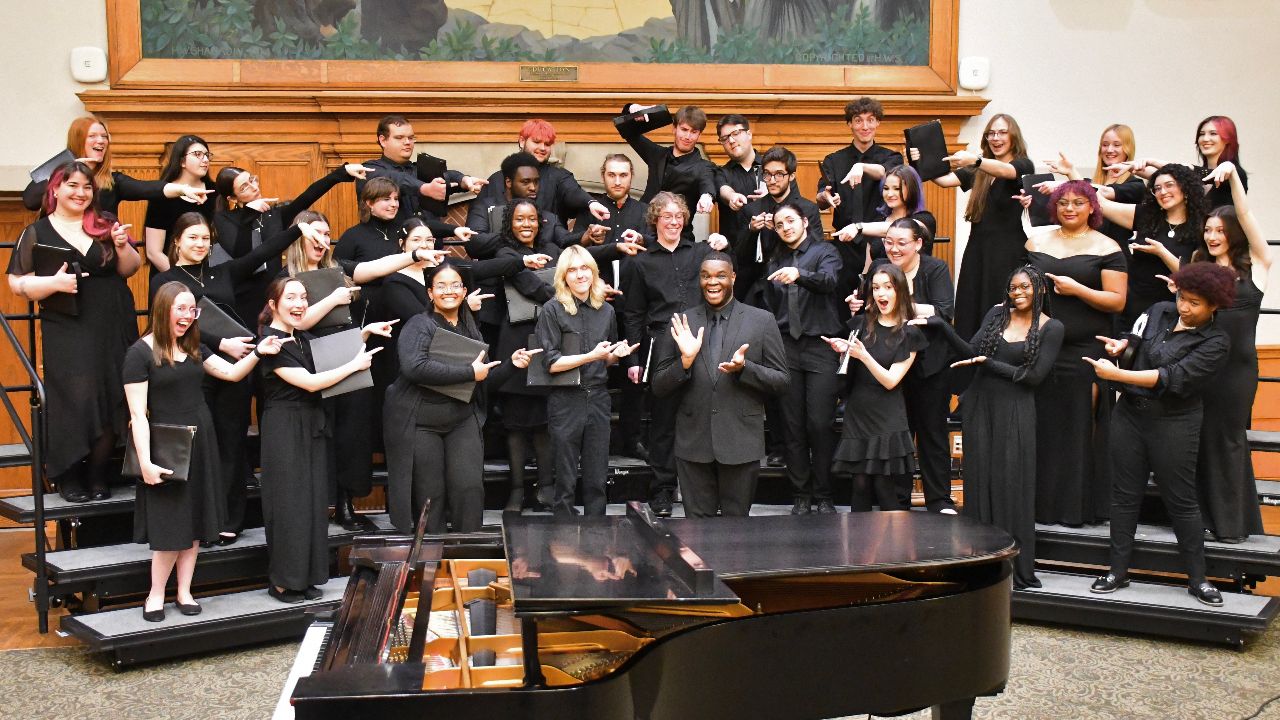A mixed chorus concert with director on risers in front of a wood background