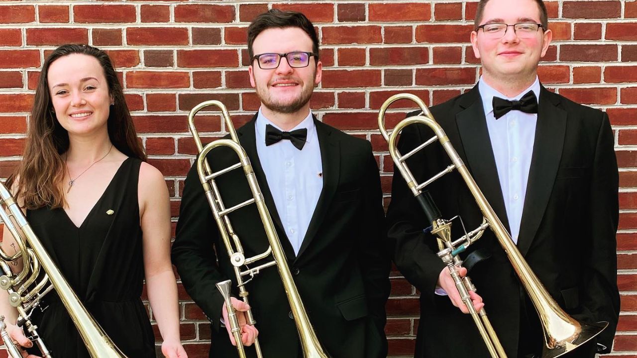3 trombone players holding their instruments in front of a brick background