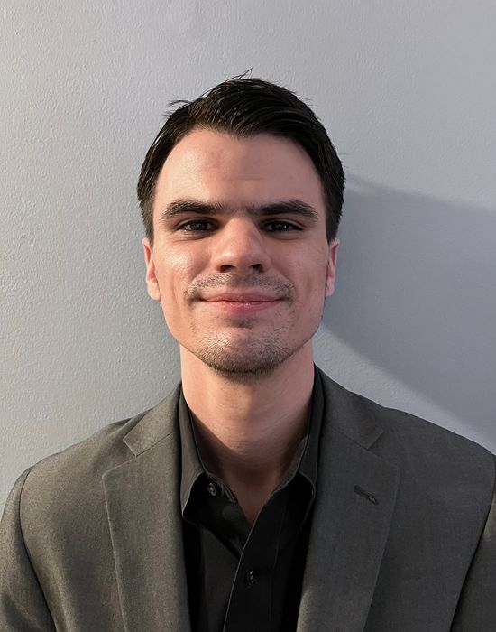 Professor's Headshot against a blueish gray wall. he is wearing a gray suit.