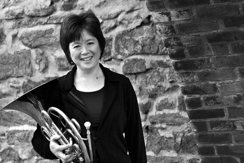 Black and white headshot of Shari Mayrhofer holding a french horn in front of a stone wall and smiling 