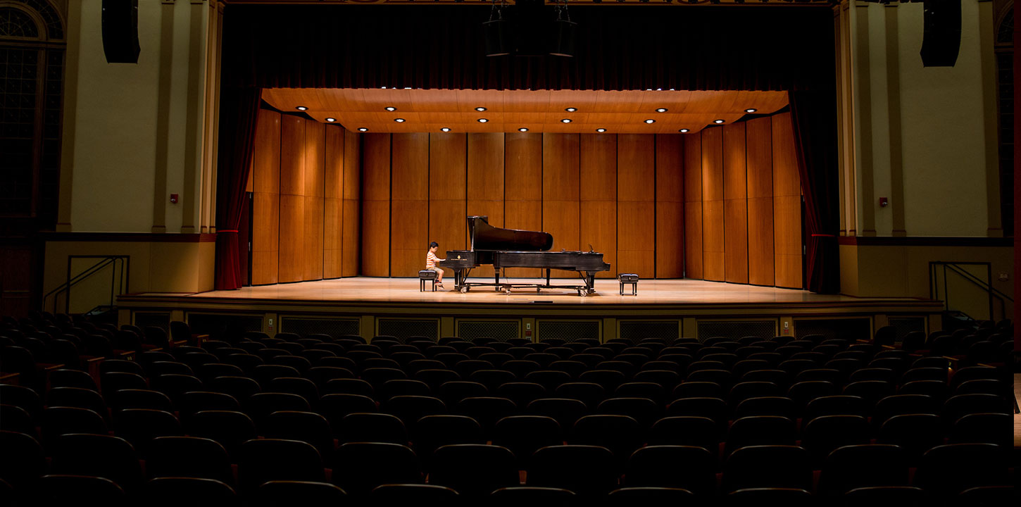 Distant shot of a student playing a grand piano on stage in an empty auditorium 