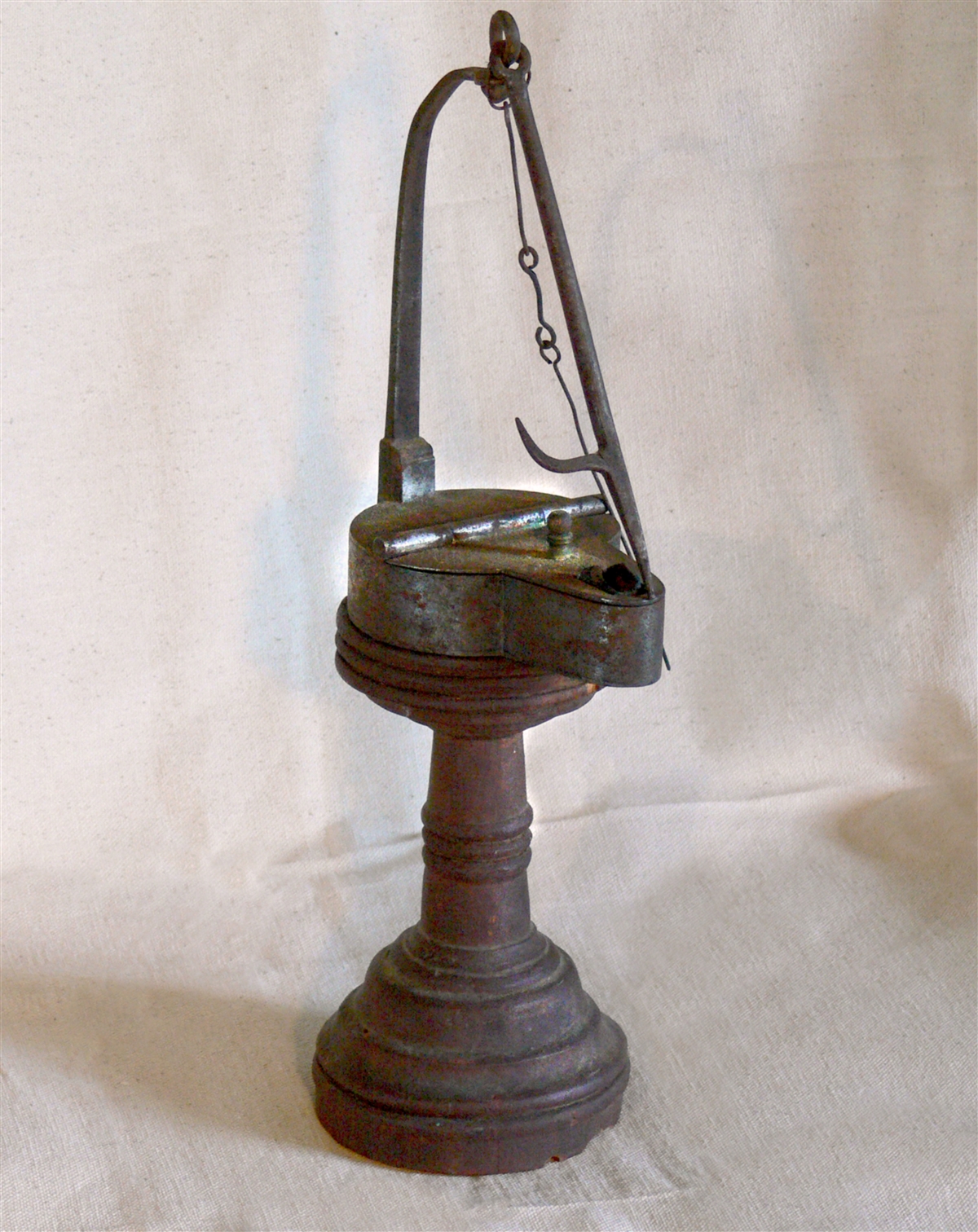 A small metal fat lamp sits on top of a wooden pedestal. The lamp is in a keyhole shape with a hinged lid. There is a small opening at the front, as well as a small metal finial. At the back is a long, curved piece of metal which holds a wick pick and anchor.