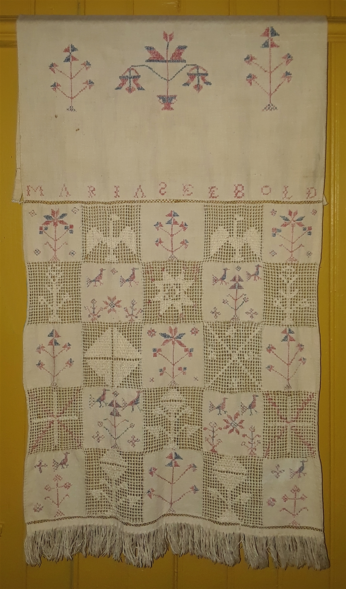 A white tea towel with pink and blue thread decorative stitching. The top portion is a solid white with a center motif of flowers in vases, with secondary floral arrangements on each side. The name Maria Seebold is stitched below. The lower portion has 12 solid fabric squares with floral arrangements stitched in pink and blue thread. 12 additional lace squares include a star, a diamond, birds, flowers, and crosses. There is a fringe at the very bottom of the tea towel.
