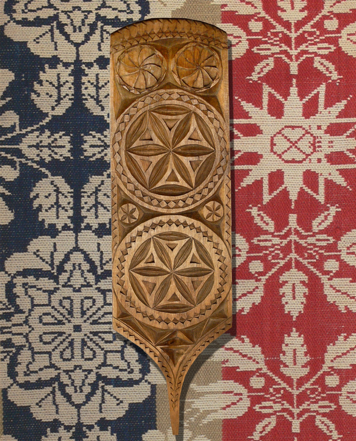 In the foreground of the image is a wooden paddle with a thin handle. The main body has two large 6-pointed chip-carved rosettes. There are two small 6-pointed rosettes between then, and two swirls at the top of the paddle. The border around the paddle is a sawtooth design. In the background is a woven coverlet. There are sections of blue, white, and red, with a repeating pattern of white stars, flowers, and leaves.