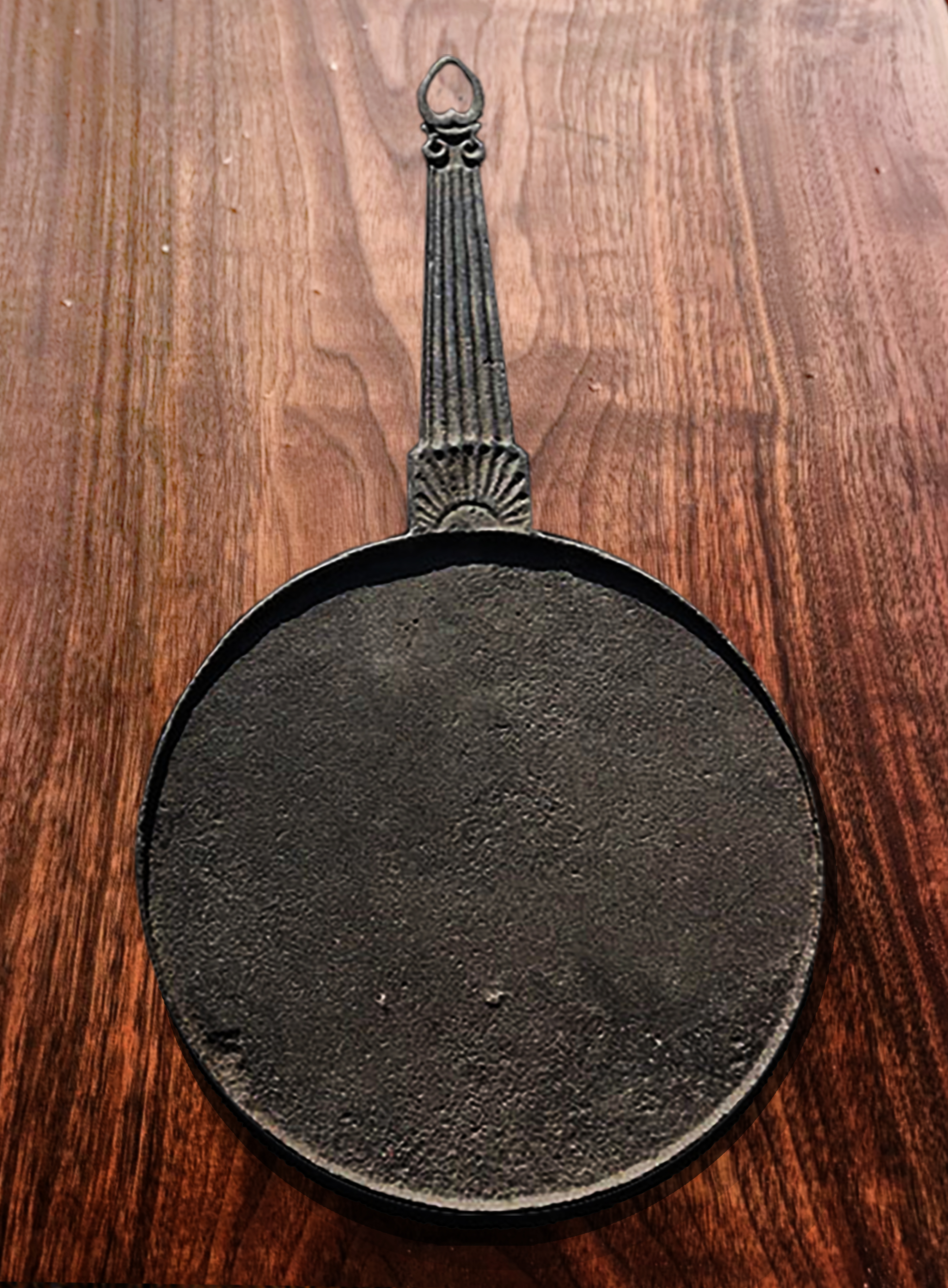 A round iron griddle with a long handle featuring a rising sun pattern at the base of the handle and a heart pattern at the tip.