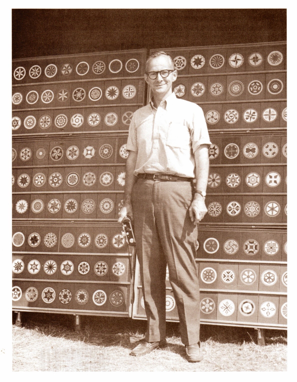 A sepia photo of a white man with white hair and dark rimmed glasses stands in front of a hex sign display. He smiles at the camera and wears a light shirt and a dark pair of pants.