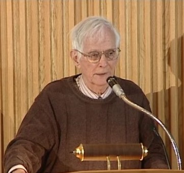 A white man with white hair and glasses stands in front of a microphone. He wears a light shirt and a brown sweater.