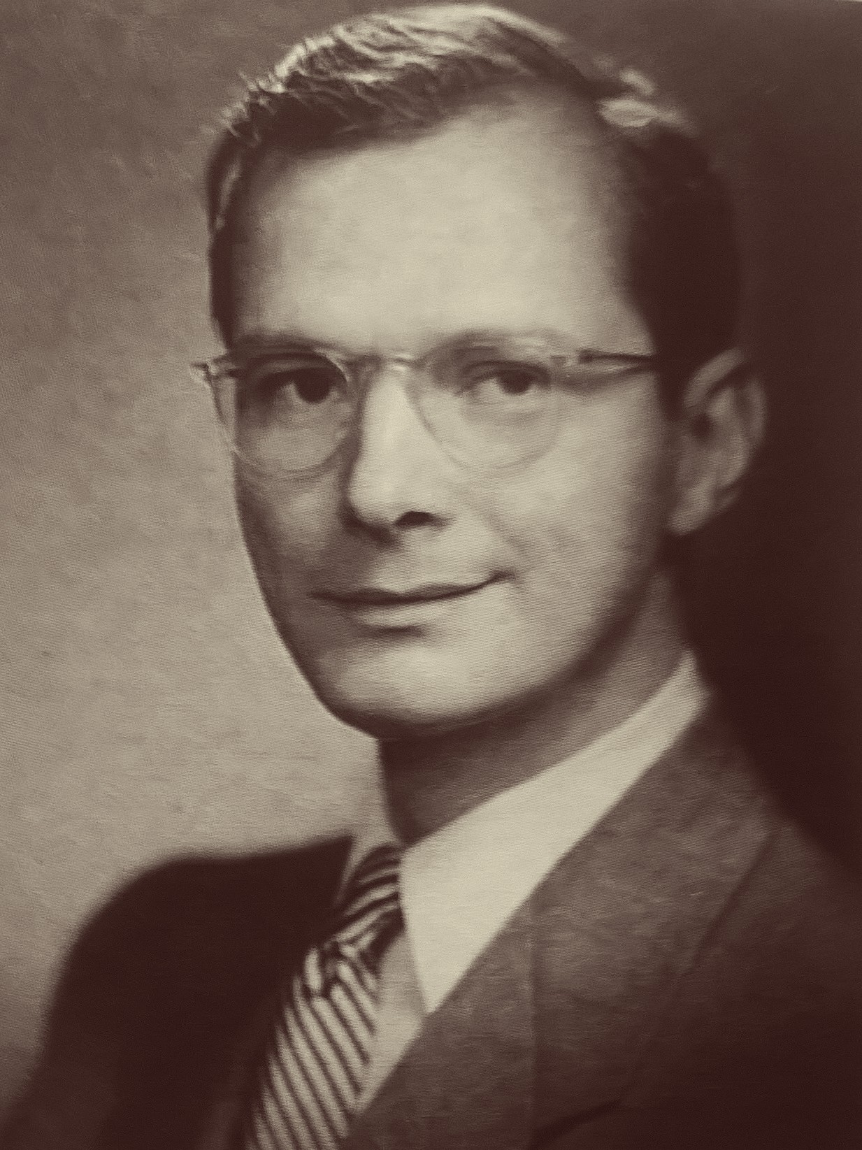 A sepia photo of a white man with dark hair smiles at the camera. He is wearing clear rimmed glasses, a light colored shirt, a striped tie, and a dark suit jacket.