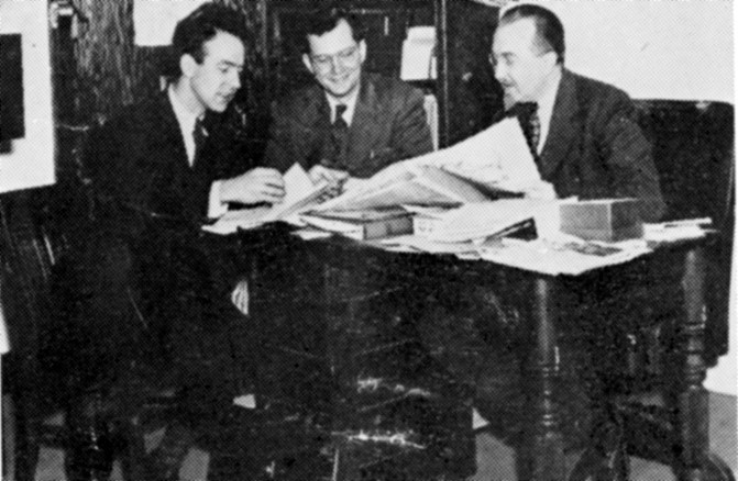 Three white men are smiling and sitting around a table. They are looking at a pile of newspapers on a table. They are smiling and wearing dark suits with light shirts.