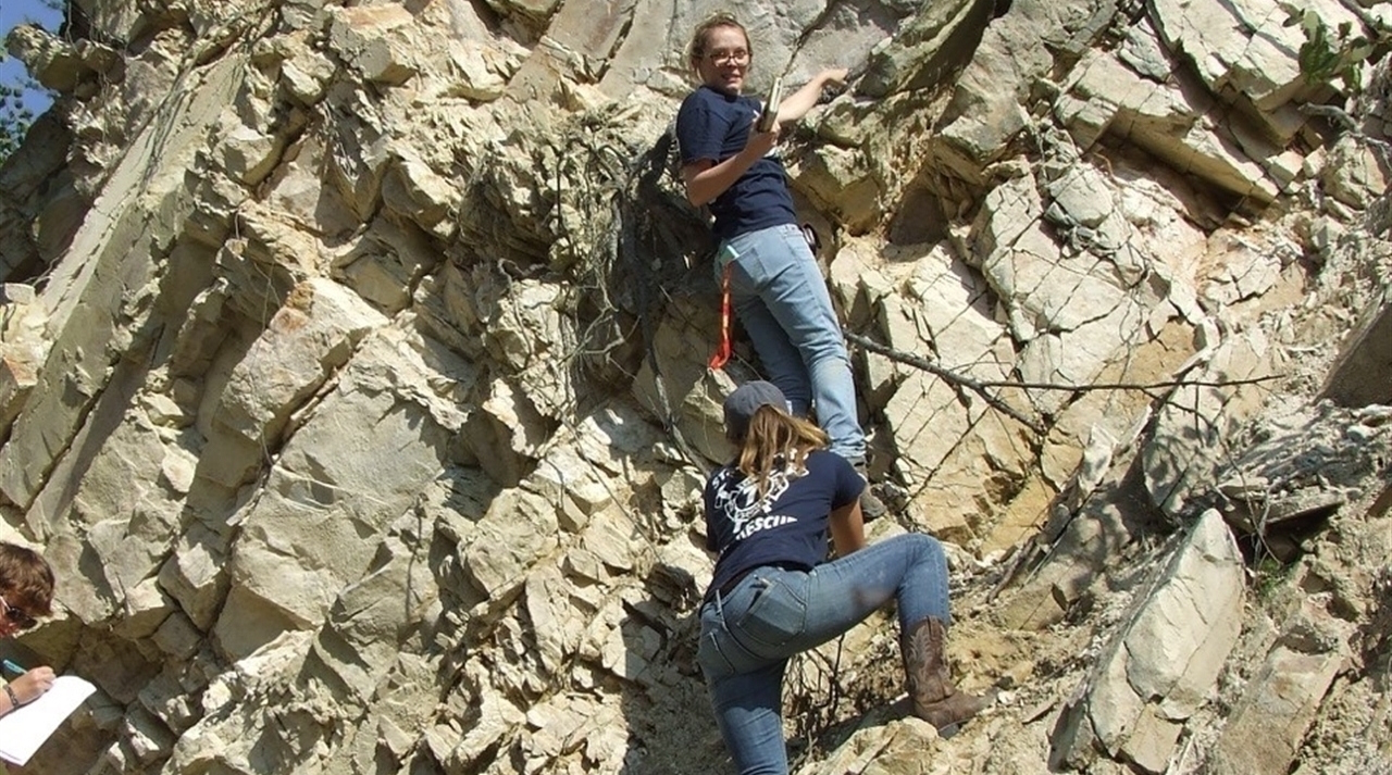 Two geology students scaling the side of a cliff, one of them looking back at the camera