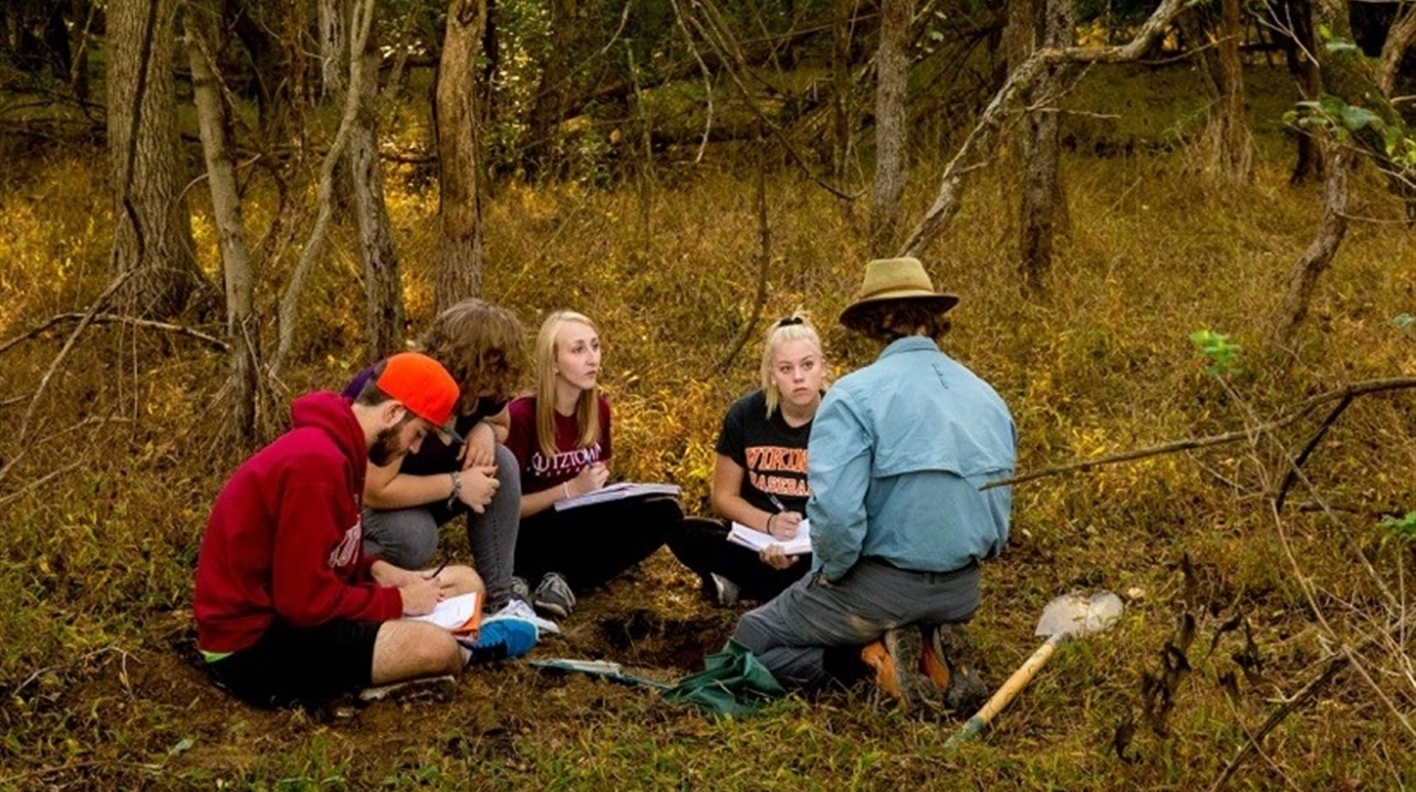 Professor lecturing to students on the forest floor as students take notes