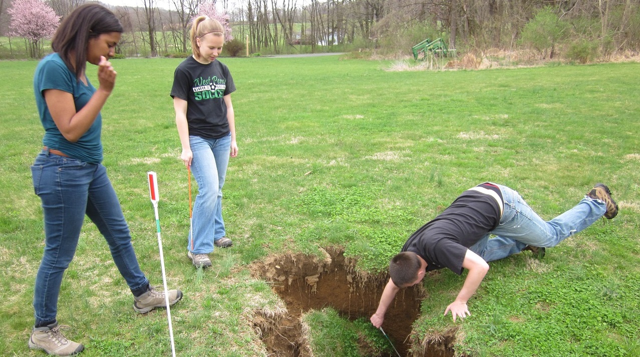 Two female students standing by while a male student reaches into a crater to gather a sample of sediment