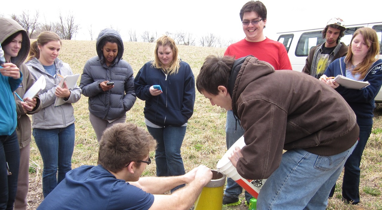 Small class standing in a circle around two of their peers, who are pouring river water into equipment for testing