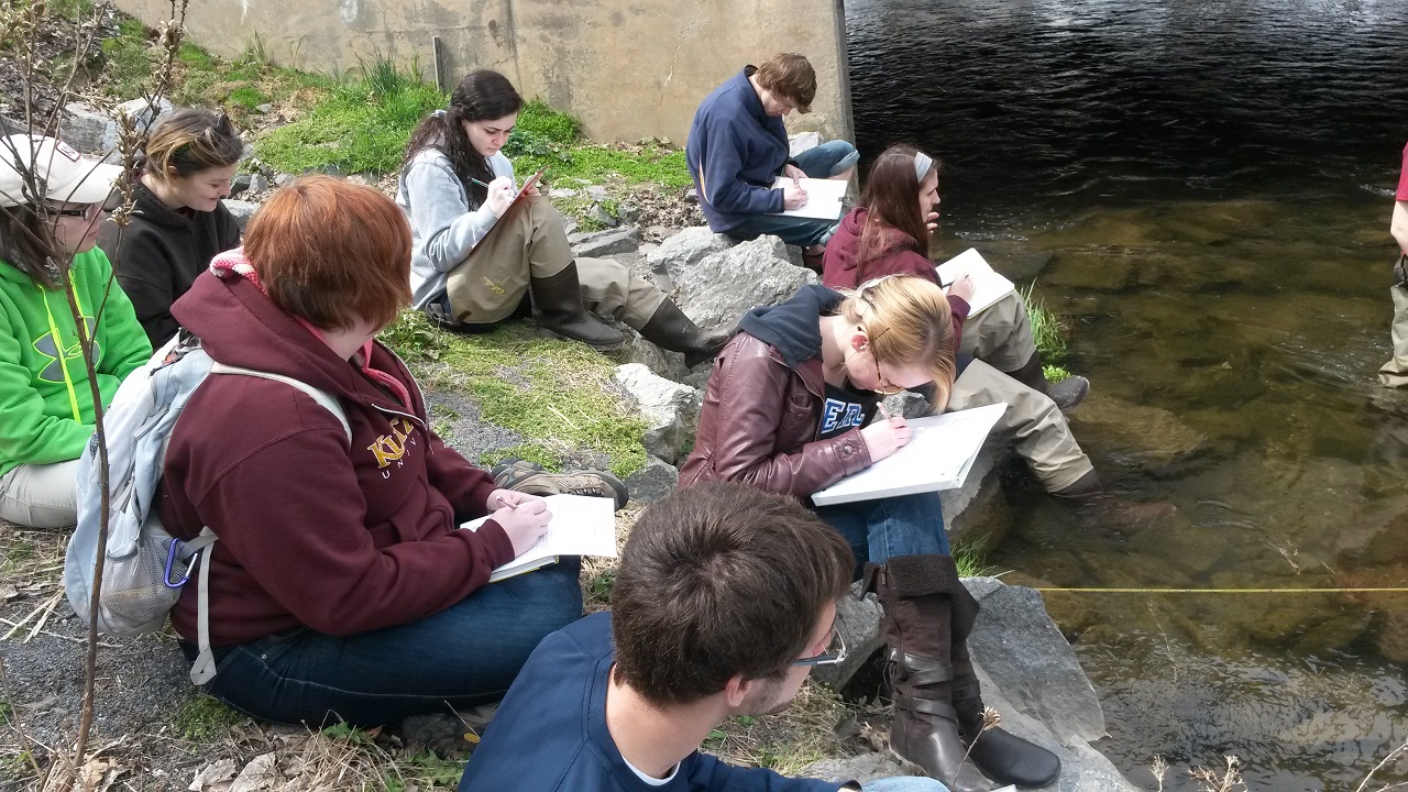 Small class of students taking notes while sitting on a riverbed 