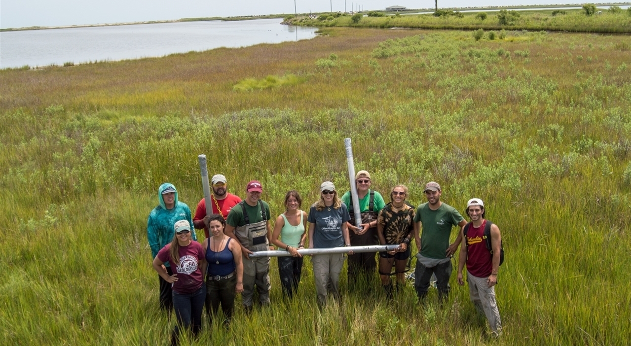 Aerial shot of geology class standing in a lakeside field, holding vibracoring equipment and smiling
