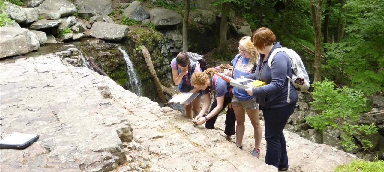 Four geology students studying rock formation. Rocks, green trees and flowing water appear in the background.