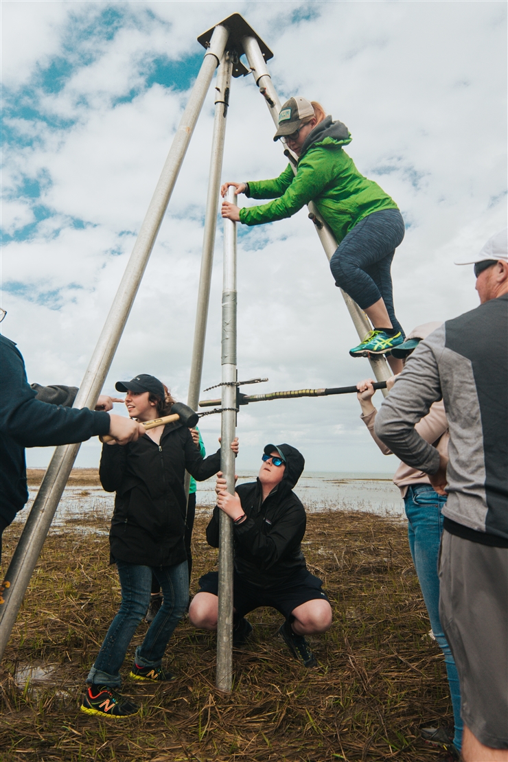 Four students hold a large tripod while another student climbs it