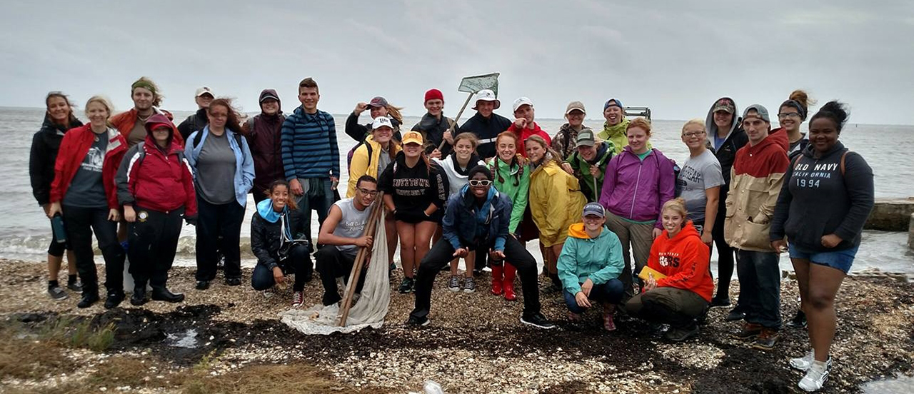 2015 Oceanography class smiling as a group on the beach, some holding nets or other oceanographer tools