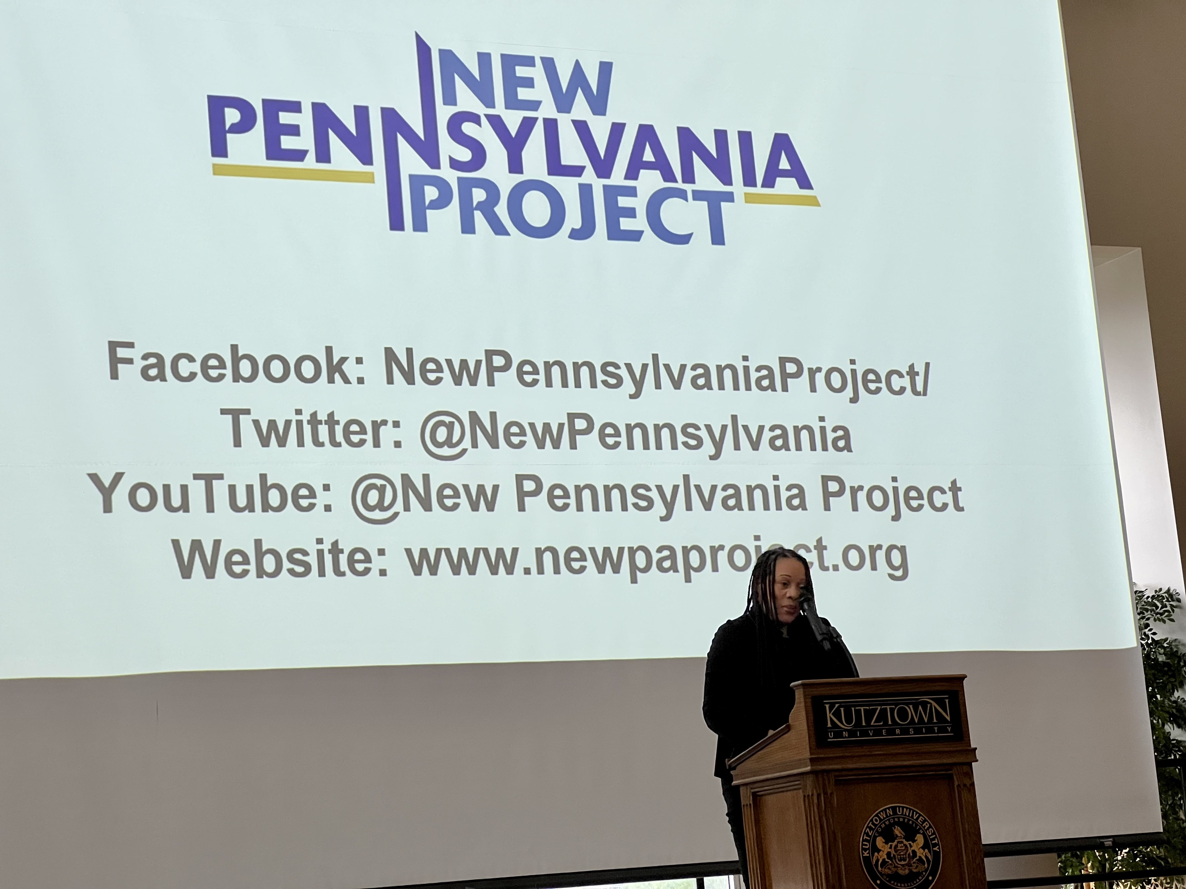 Person standing at KU podium with screen projecting New Pennsylvania Project behind