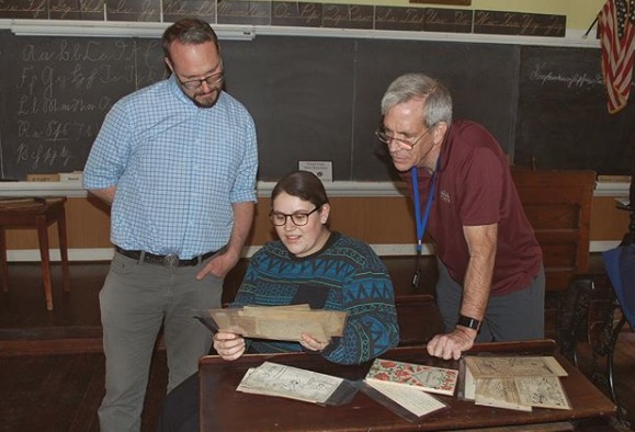 Two professors stand on either side of a student, all of them analyzing a browned page