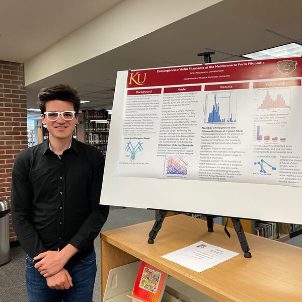 Student in Black formal attire standing next to his KU Inspires Poster
