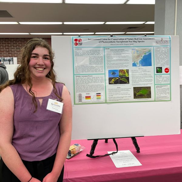 Student wearing purple blouse stands in front of her poster and smiles