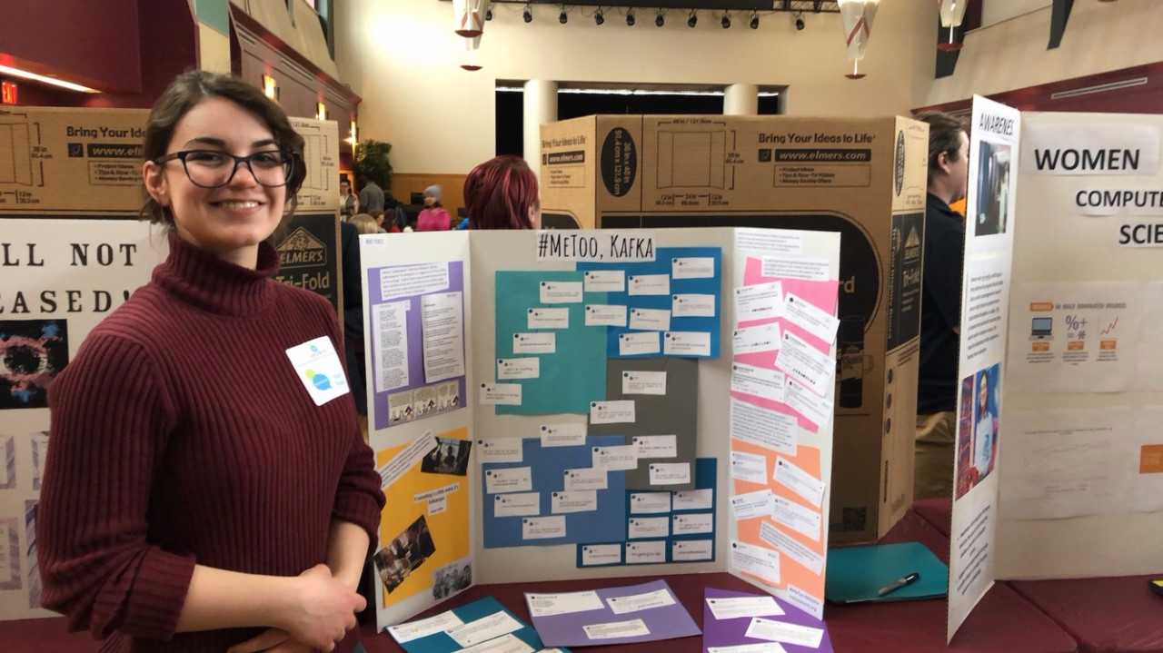 Female student Noel Fedeli smiling while standing beside a tri-fold titled "#Me too, Kafka" at KUs Council on Human Diversity Conference
