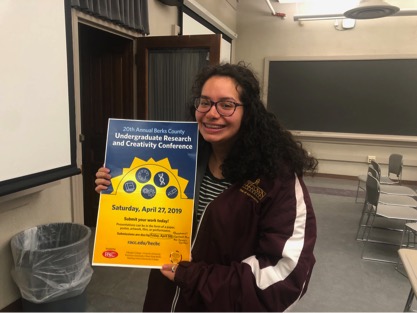 Female student smiling and holding up a poster for the undergraduate research and creativity conference