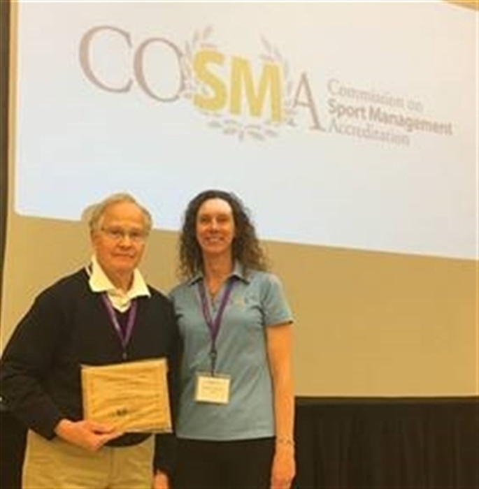 professor standing on stage and holding his inductee award on stage next to a female society member, in front of a COSMA banner