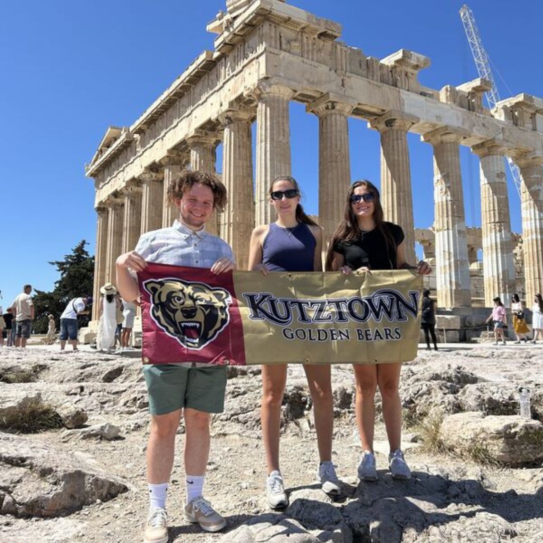 Three Sport Management students holding a Kutztown University Golden Bears banner in front of the Acropolis in Athens, Greece.