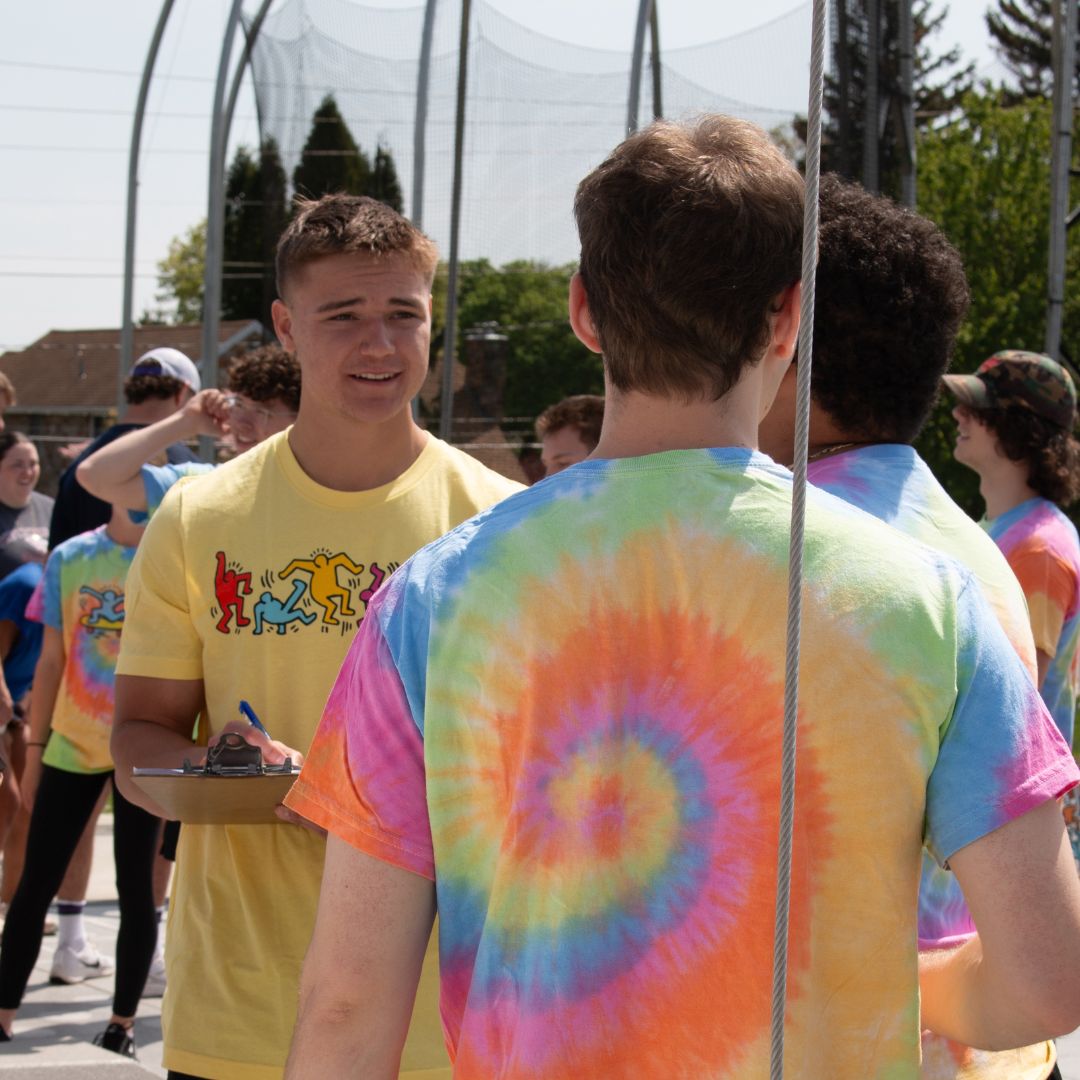 A Sport Management student in a yellow Keith Haring T-shirt holds a clipboard while speaking with peers in tie-dye shirts during an outdoor activity.
