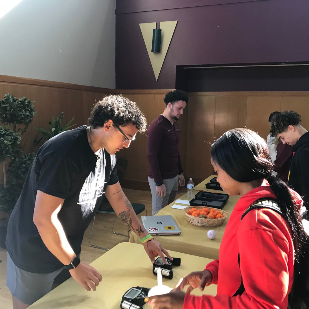 A Sport Management student demonstrates equipment to a visitor at a table during a Wellness Expo, with other students engaging participants in the background.