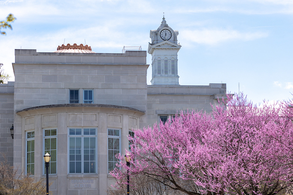 The clock tower of Old Main peeks out to the right of the Graduate Center dome with a pink flowering tree in Spring in front.