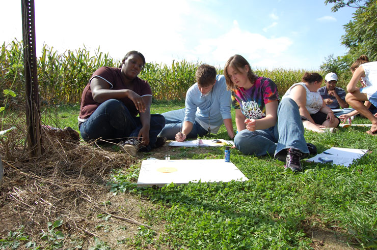 A photo of students sitting on the grass next to a corn field doing academic work at KU's eco-plot. 
